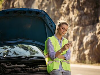 A woman with a lighted vest looks into her cell phone and waits for roadside assistance. She leans against her black car.