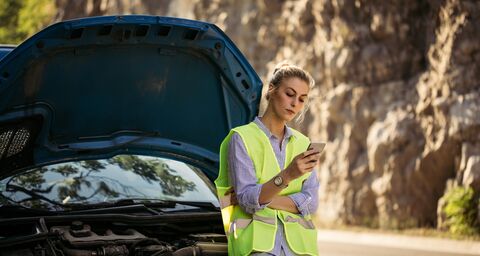 Una donna con un giubbino catarifrangente guarda nel suo cellulare e aspetta il soccorso stradale. Si appoggia alla sua auto nera.