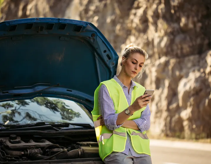 A woman with a lighted vest looks into her cell phone and waits for roadside assistance. She leans against her black car.