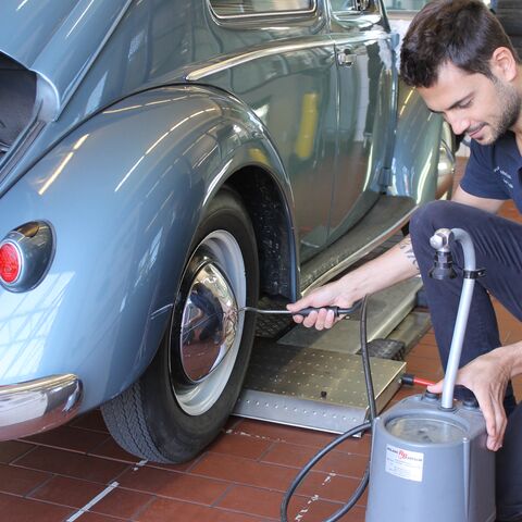 Un uomo controlla la pressione degli pneumatici di un'auto d'epoca blu in un'officina, utilizzando un dispositivo portatile per la pressione dell'aria.