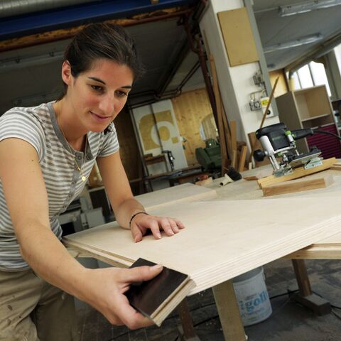 Eine Frau in einem Holzarbeitsraum arbeitet konzentriert an einem Stück Holz, umgeben von Werkzeugen und Maschinen, mit einem Fenster im Hintergrund.