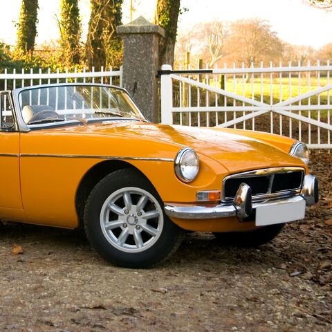 Un'auto sportiva gialla e classica decappottabile è parcheggiata su una pista sterrata davanti a una recinzione bianca e ad alberi sullo sfondo.