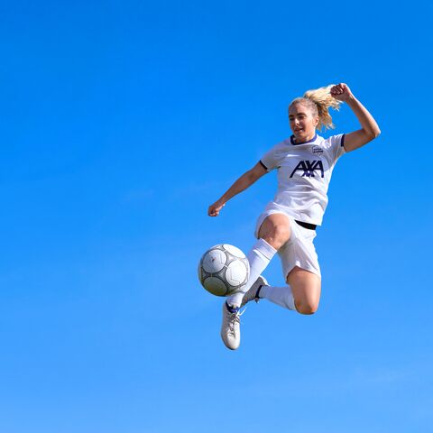 Una calciatrice con la maglietta bianca AXA salta in aria e calcia un pallone da calcio davanti a un cielo terso.