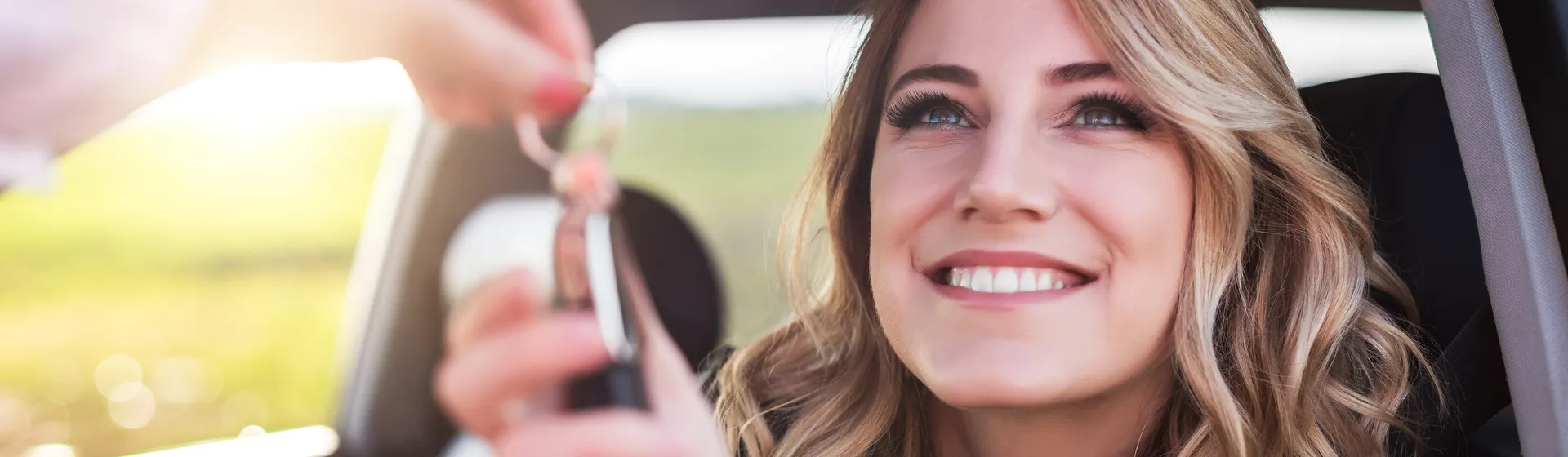 A smiling woman with long hair is sitting in a car and receives a car key from another person through the open window.