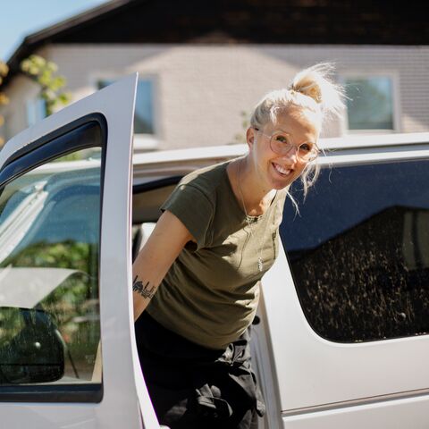 Junge Frau mit blonden Haaren, Brille und grünem T-Shirt steigt lachend aus einem weissen Auto.