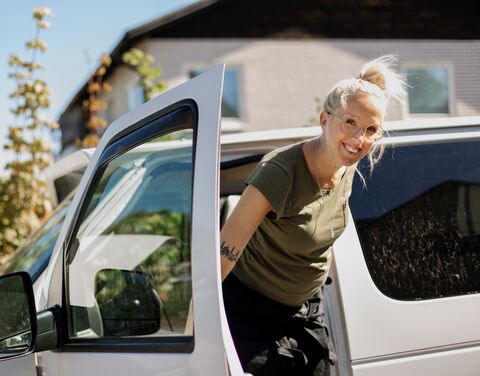 Junge Frau mit blonden Haaren, Brille und grünem T-Shirt steigt lachend aus einem weissen Auto.