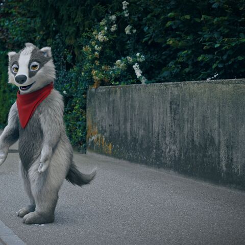 Max le futé vêtu de son foulard rouge est sur le trottoir d’une rue d’un faubourg, devant une haie verdoyante.