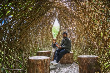 A father sits with his son on tree stumps in a willow hut.