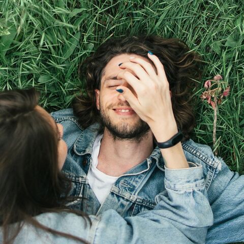 A man with long hair lies smiling in the grass while a woman gently touches his face. Both are wearing denim jackets.