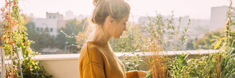 Eine Frau steht in der Sonne auf ihrem Balkon und kümmert sich um ihre Pflanzen.