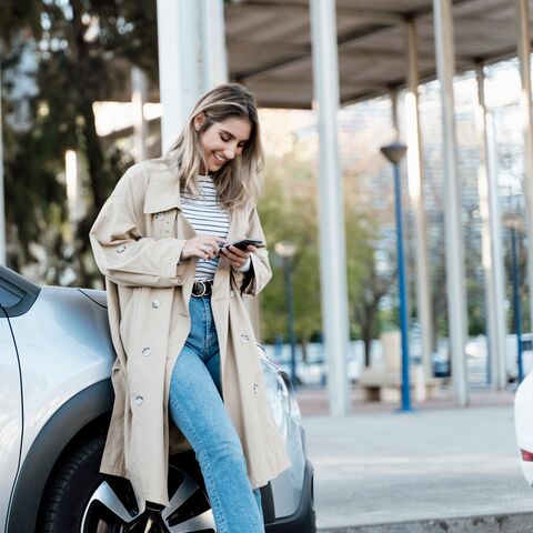 Eine junge Frau lehnt lächelnd an einem Auto, trägt einen beigen Trenchcoat und Jeans und schaut auf ihr Smartphone, während sie draussen steht.
