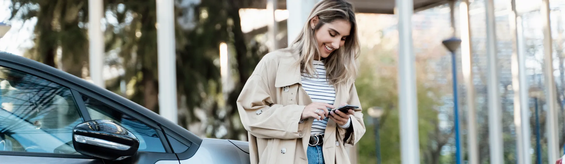 Eine junge Frau lehnt lächelnd an einem Auto, trägt einen beigen Trenchcoat und Jeans und schaut auf ihr Smartphone, während sie draussen steht.
