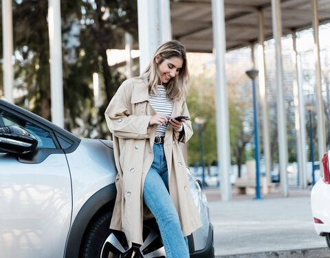Eine junge Frau lehnt lächelnd an einem Auto, trägt einen beigen Trenchcoat und Jeans und schaut auf ihr Smartphone, während sie draussen steht.