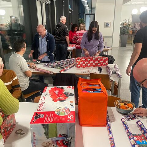 Several people wrap presents together at tables in a bright room.