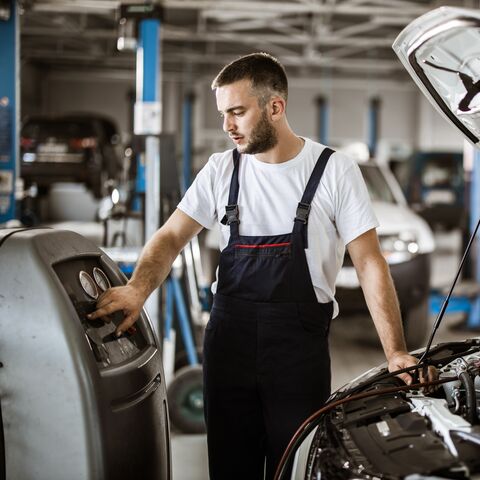 Ein Mechaniker in Arbeitskleidung überprüft ein Diagnosegerät neben einem Auto mit offener Motorhaube in einer Werkstatt.