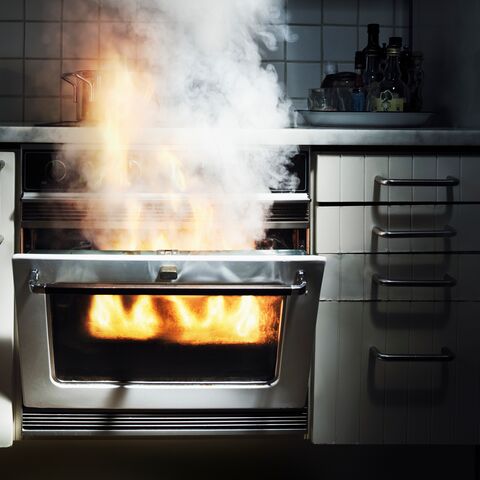 Un forno aperto in una cucina da cui emergono fiamme e fumo denso, circondato da armadi bianchi.