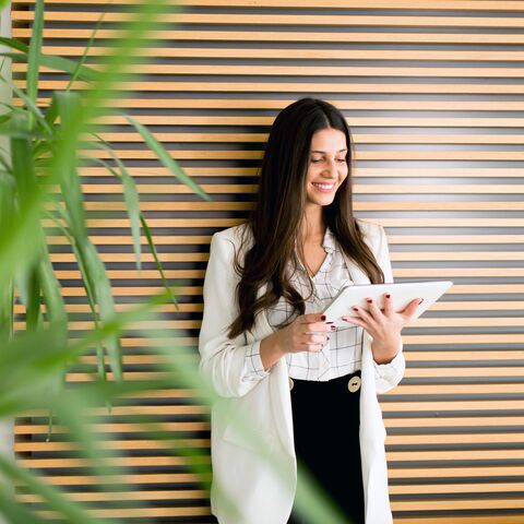 Une femme souriante vêtue d'un blazer blanc se tient devant un mur de lamelles de bois et tient une tablette ; à gauche, des plantes vertes au premier plan.