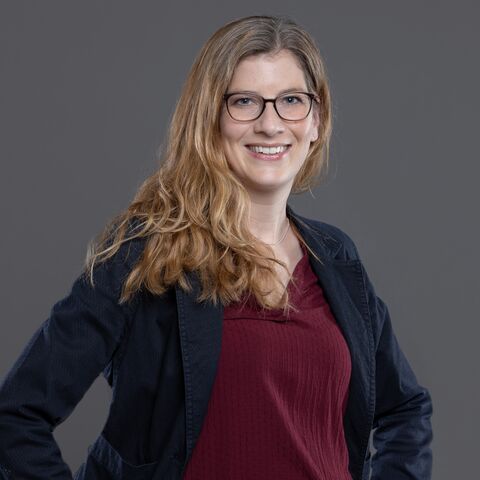 Una giovane donna con i capelli rossicci e gli occhiali sorride, sicura di sé, alla telecamera. Ha le mani sui fianchi e indossa un blazer blu scuro e una camicetta di color bordeaux.