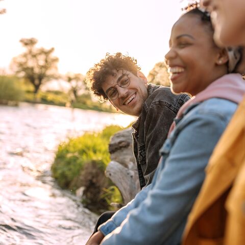 Trois amis souriants sont assis au bord de la rivière, profitent du soleil du soir et discutent joyeusement, tandis que la nature s’épanouit en arrière-plan.