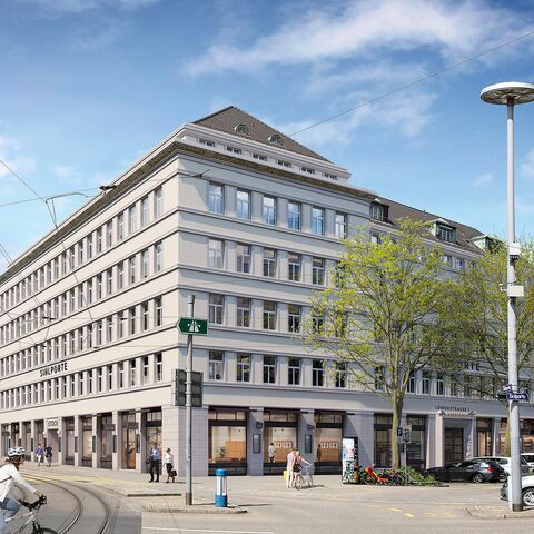 Ein mehrstöckiges, klassisches Gebäude an einer Strassenecke mit Geschäften im Erdgeschoss, Bäumen und Menschen auf der Strasse bei sonnigem Wetter.