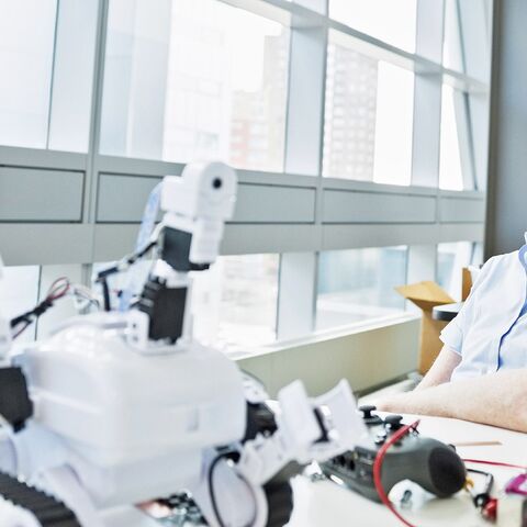 Ein Mann mit Brille sitzt an einem Schreibtisch und betrachtet einen weissen Roboter, umgeben von Elektronik und Werkzeugen, vor einem grossen Fenster.