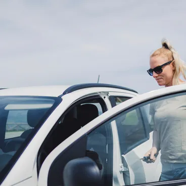 Eine blonde Frau mit Sonnenbrille öffnet die Tür eines weissen Autos und hält den Autoschlüssel in der Hand.
