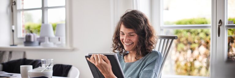 Una donna siede a un tavolo in una stanza luminosa e guarda, sorridendo, lo schermo di un tablet.