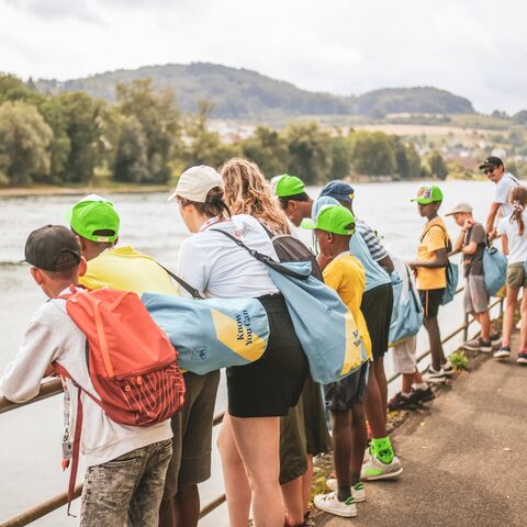 A group of children and adults stand on a sidewalk on a riverbank and look curiously over the railing into the water.
