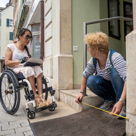 A woman in a wheelchair and another person measure the height of an entrance step in a busy pedestrian zone together.