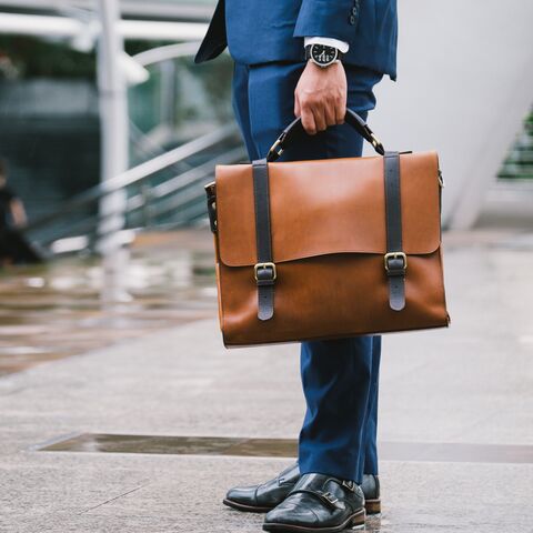 A man in a blue suit holds a brown leather bag and stands on a wet sidewalk in front of a modern urban background.