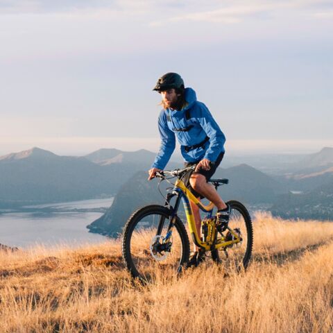 Un vététiste vêtu d’un casque noir et d’une veste bleue roule sur une crête. En arrière-plan, on voit un lac entouré de chaînes de montagnes.