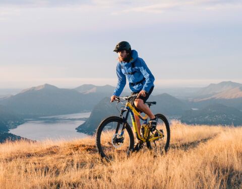 Man in a blue jacket and bike helmet is riding a yellow mountain bike. In the background we see the surrounding mountains and a lake in the valley.