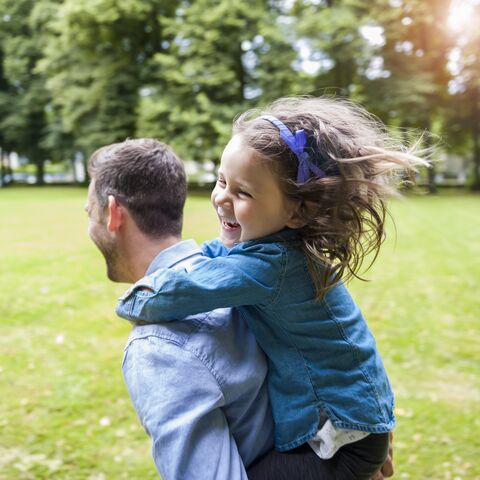 Un uomo porta sulle spalle una ragazza ridente dai capelli fluenti attraverso un parco verde in una giornata di sole.