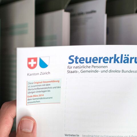 Une main tient une déclaration d’impôts du canton de Zurich sur un fond flou avec des étagères remplies de classeurs.