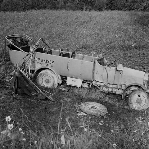 A badly damaged old car lies abandoned in a meadow. A tire and debris are scattered around the vehicle.