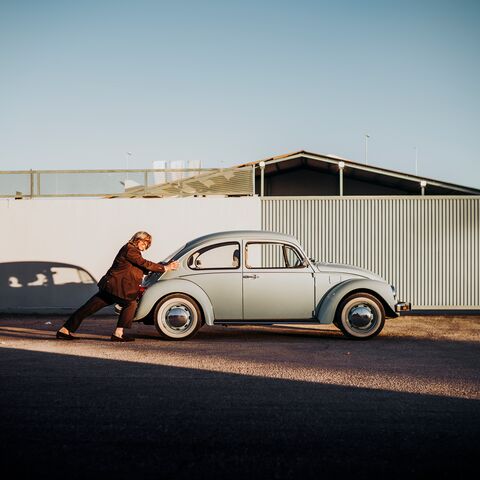 Eine Person schiebt einen hellblauen, klassischen VW Käfer entlang einer Strasse, die von einer grauen Wand und einem Gebäude gesäumt ist.