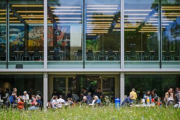 Un bâtiment moderne avec une grande façade en verre, à travers laquelle on peut voir un intérieur lumineux avec des tables et des chaises. En dessous, de nombreuses personnes sont assises devant une pelouse verte.