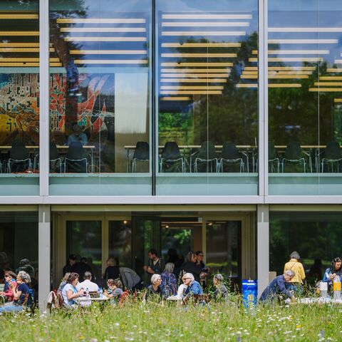 A modern building with a large glass façade through which a bright interior with tables and chairs can be seen. Underneath, many people sit on a green lawn