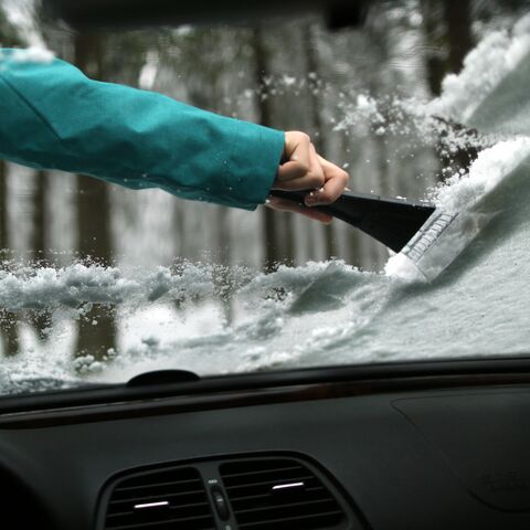 Una persona con una giacca turchese rimuove con un raschiaghiaccio la neve dal parabrezza di un’auto in un bosco invernale.
