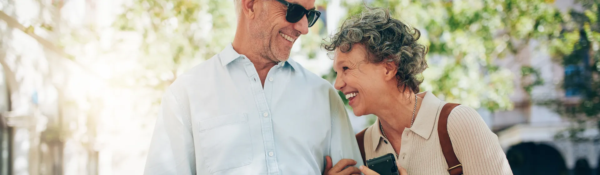 Una coppia di anziani sorridenti all’aperto: lei tiene in mano uno smartphone mentre lui legge una cartina; entrambi indossano abiti estivi casual.