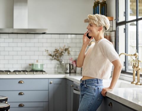A young blonde woman is at home in the kitchen talking on the phone.