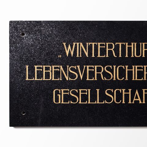 Black sign with gold lettering shows the text “WINTERTHUR LEBENSVERSICHERUNGS GESELLSCHAFT” on a white background.