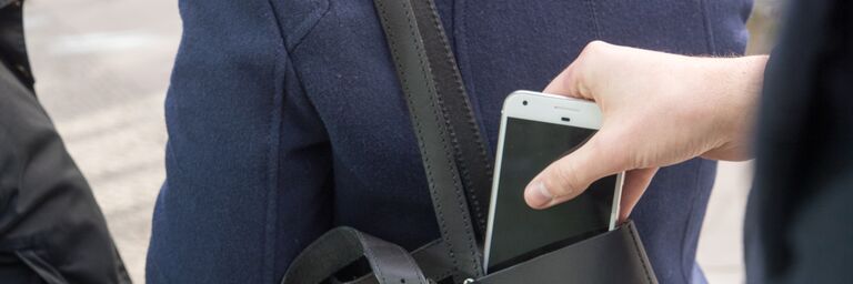 Eine Person entwendet ein Mobiltelefon aus einer umgehängten Handtasche.