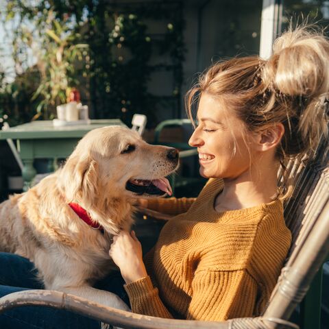 A woman sits outside with a smile, caressing a Golden Retriever, who looks at her affectionately; in the background are plants and a table.