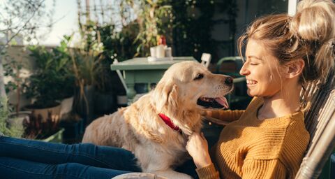 Une femme est assise dehors en souriant et caresse un Golden Retriever qui la regarde affectueusement; à l’arrière-plan, il y a des plantes et une table.