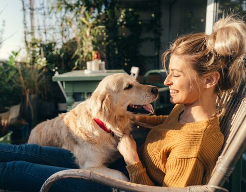 Eine Frau sitzt lächelnd draussen und streichelt einen Golden Retriever, der sie liebevoll ansieht; im Hintergrund sind Pflanzen und ein Tisch.