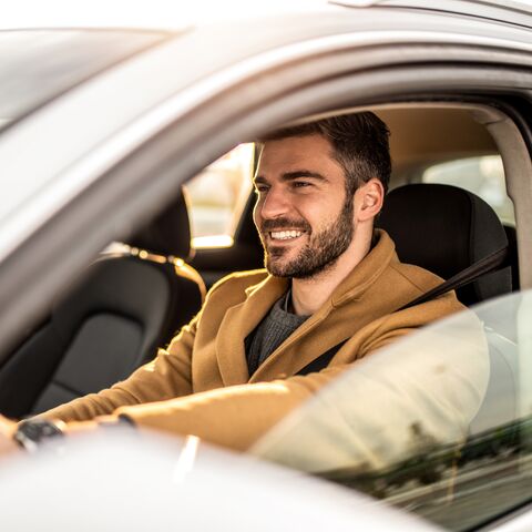 Ein Mann mit Bart fährt lächelnd in seinem Auto weg.