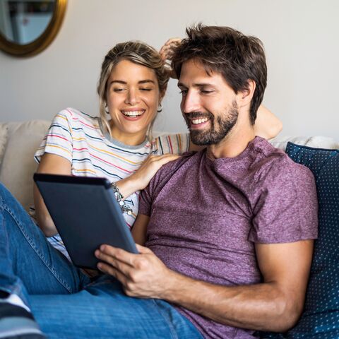 A young couple is sitting on the sofa at home. Both are looking at the tablet the man is holding in his hands.