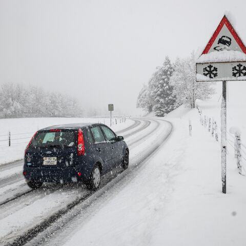 Une voiture roule sur une route de campagne enneigée, entourée d’arbres enneigés; un panneau de signalisation signale qu’il y a de la neige et du verglas.
