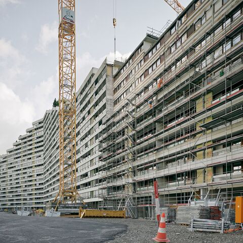 Eine grosse Baustelle mit hohen, modern designten Wohngebäuden im Hintergrund, eingerüstet und von einem gelben Kran überragt.